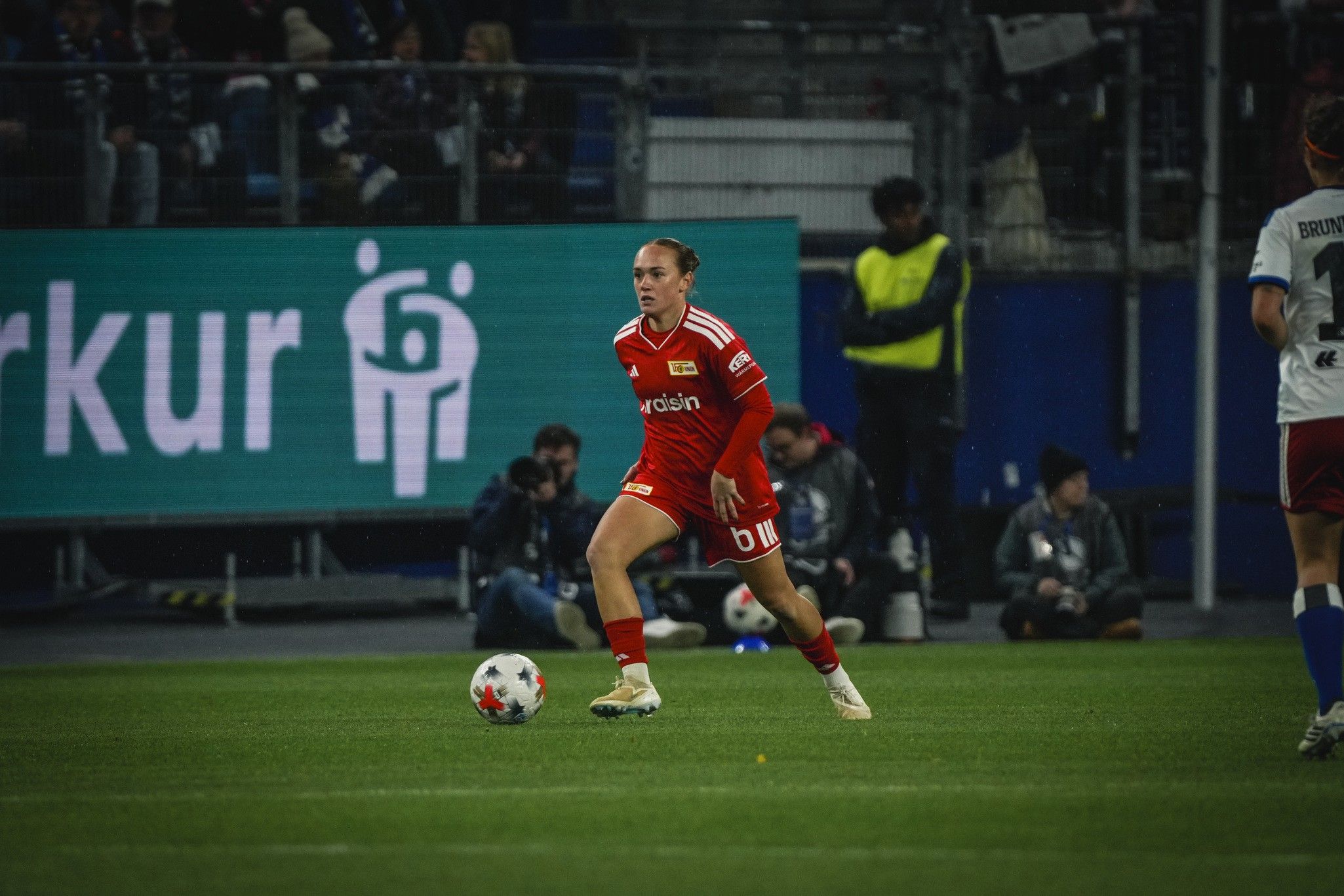 Eine Fußballspielerin in rotem Trikot dribbelt mit dem Ball auf einem Spielfeld, während Zuschauer und Fotografen im Hintergrund zu sehen sind.
