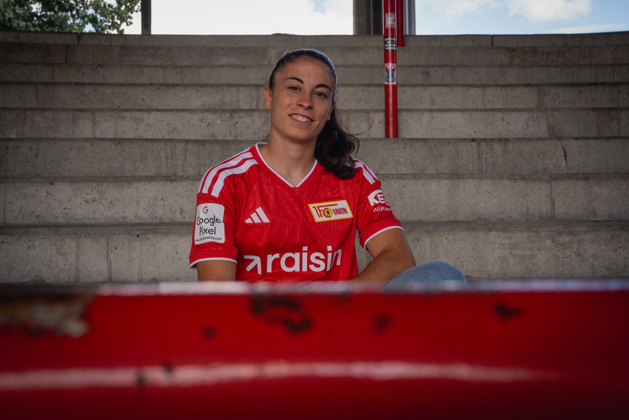 Eine Frau im roten Trikot sitzt lächelnd auf einer Treppe.