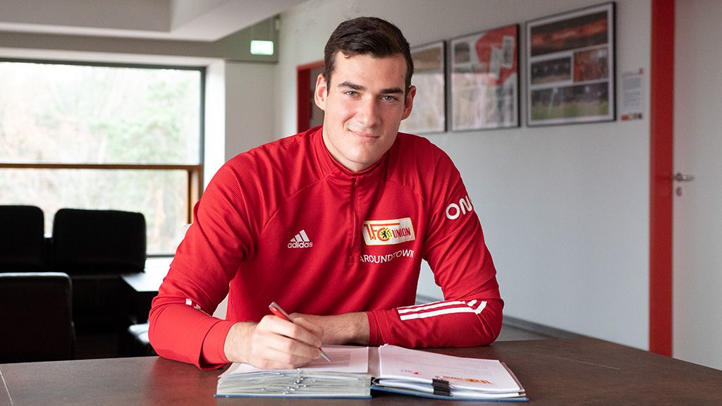 A young man in a red tracksuit sits at a table writing in a notebook. Pictures are on the wall in the background.