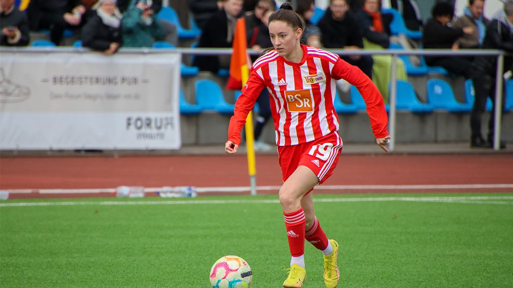 Player of FC Union Berlin in a red jersey with white stripes is dribbling the ball on a soccer field.
