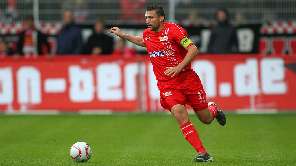 A soccer player in a red jersey sprints across the field while spectators stand in the background.