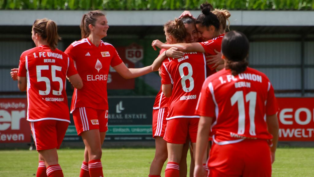 Frauenfußballmannschaft in roten Trikots feiert einen Torerfolg auf dem Spielfeld. Spielerinnen umarmen sich.
