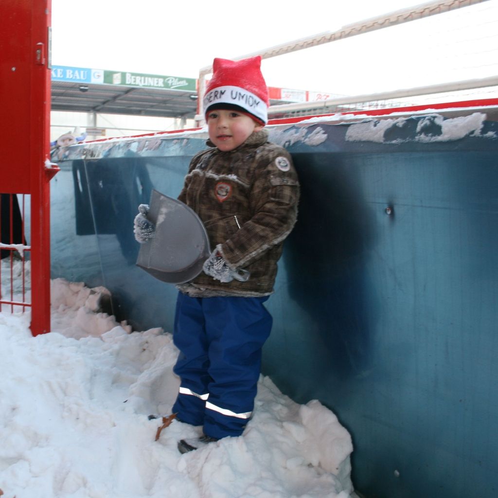 Ein Junge im Schnee trägt einen Weihnachtsmannhut und hält eine Schaufel. Er steht an einer blauen Wand.
