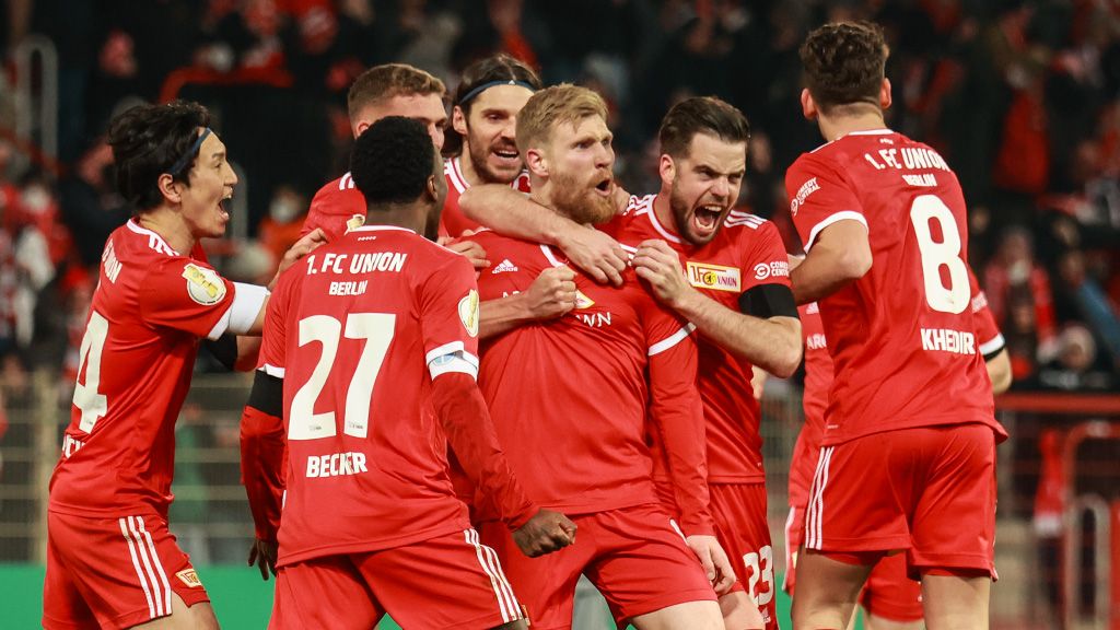Die Spieler von 1. FC Union Berlin freuen sich über ein Tor. Sie stehen auf dem Feld. Ihre Teamkollegen in roten Trikots jubeln.