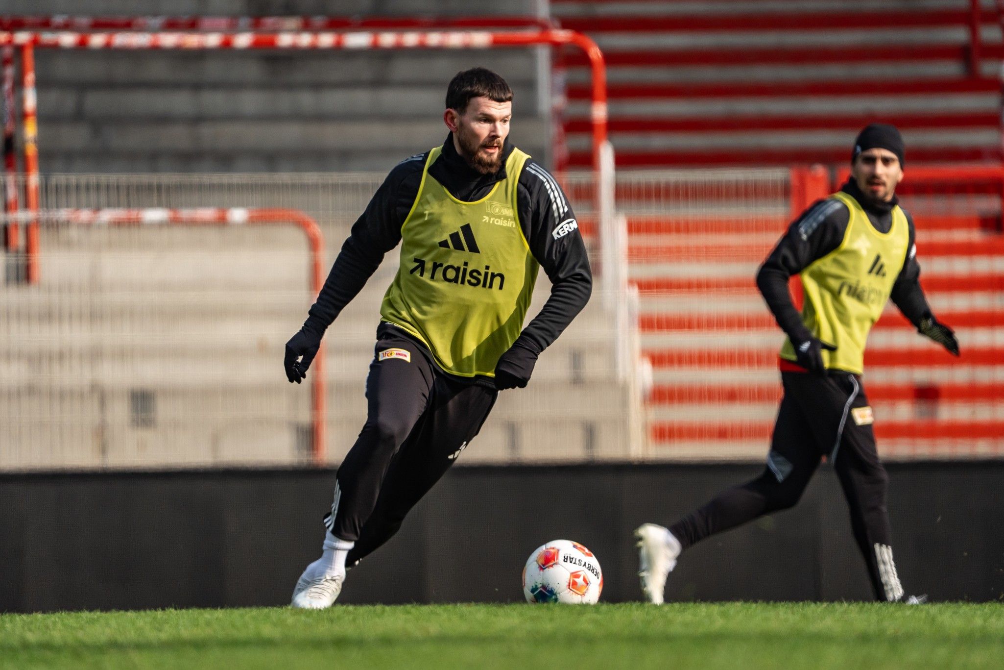 Zwei Fußballspieler in Trainingsanzügen dribbeln einen Ball auf einem Fußballfeld, umgeben von leeren Tribünen.
