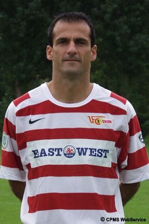 Ein Fußballspieler trägt ein rot-weiß gestreiftes Trikot mit dem Schriftzug "EAST WEST" und steht im Freien.