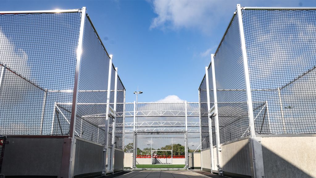 Offene Tore zu einem Sportplatz, umgeben von hohen Netzen und unter einem klaren blauen Himmel.
