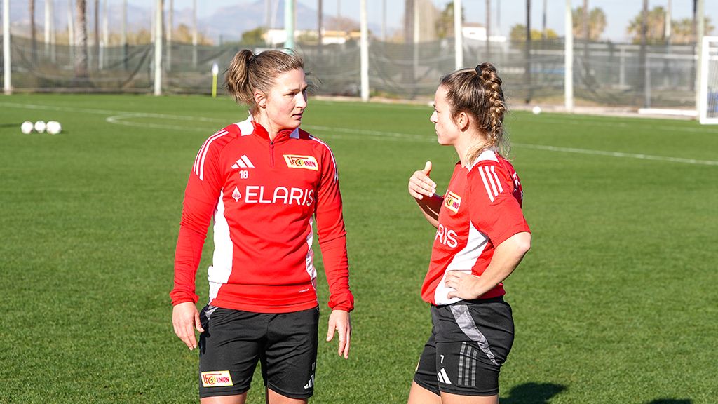 Two female soccer players in red jerseys are talking on a training field.
