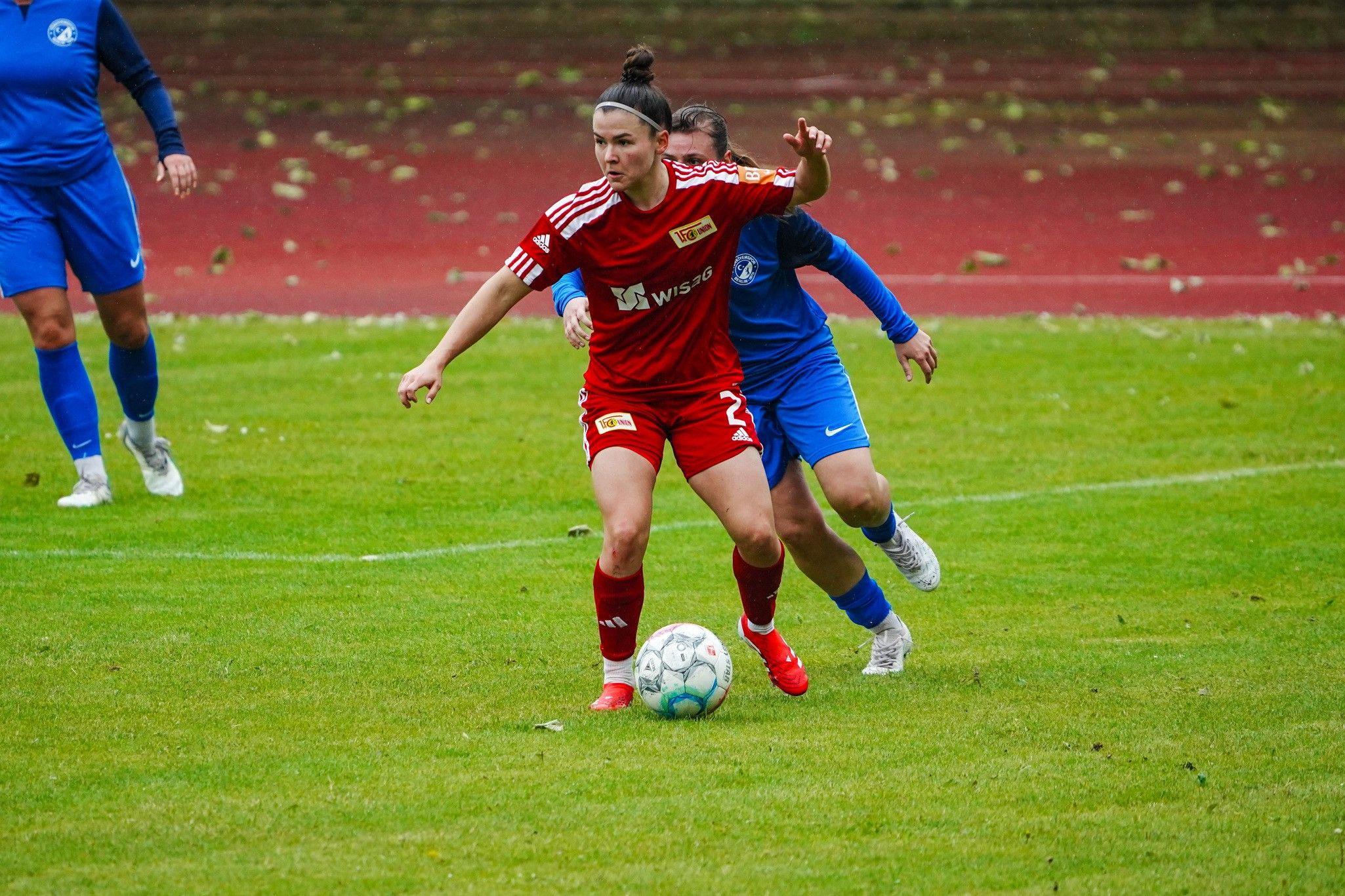 Eine Fußballspielerin in rotem Trikot dribbelt den Ball, während eine gegnerische Spielerin in blau versucht, den Ball zu erobern.