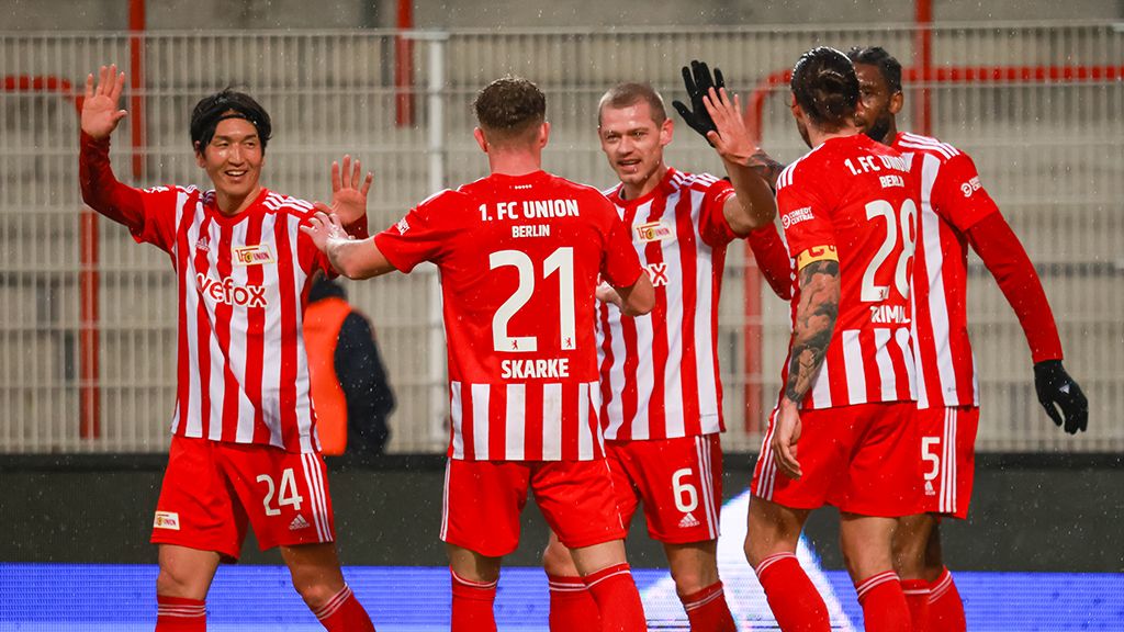 Spieler vom 1. FC Union Berlin freuen sich. Sie haben ein Tor geschossen. Sie tragen rot gestreifte Trikots. Sie sind im Stadion.