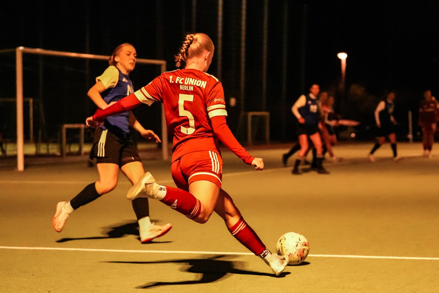 Eine Spielerin in rotem Trikot schießt den Ball auf einem Fußballplatz bei Nacht, während eine Gegenspielerin in Blau steht.