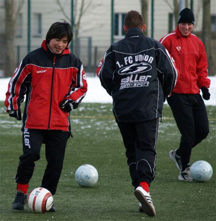 Drei Fußballspieler in Trainingsanzügen auf einem Rasenplatz, umgeben von Bällen und Schnee.