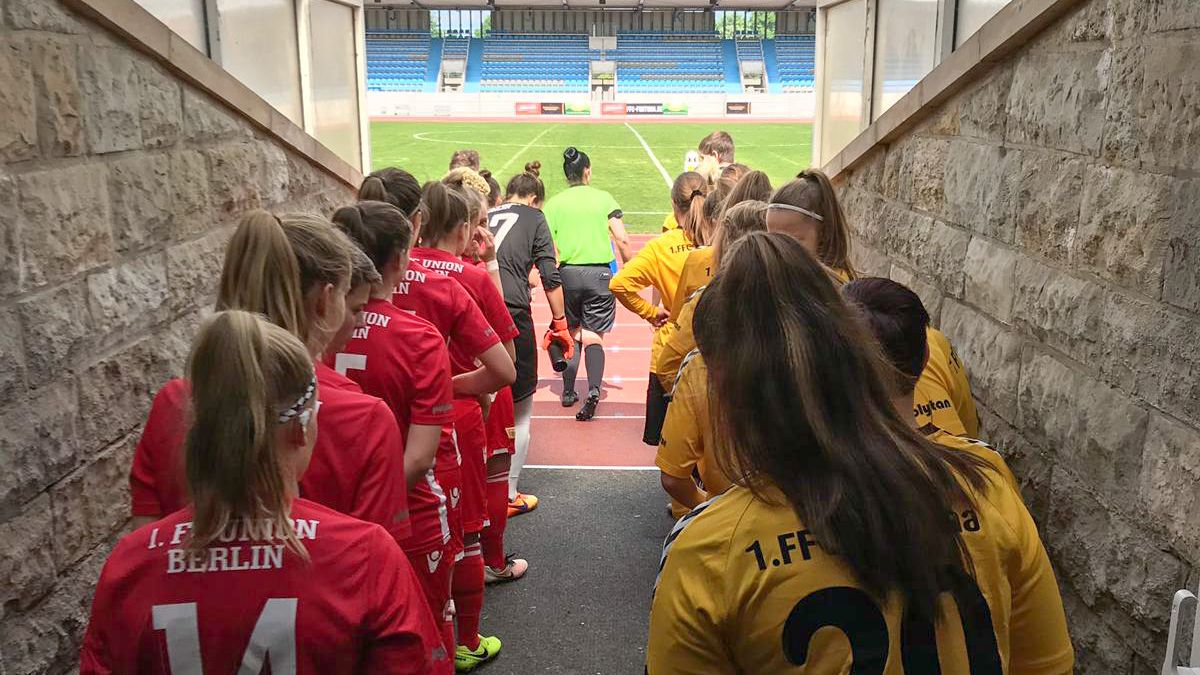Spielerinnen in roten und gelben Trikots betreten das Stadionfeld durch den Tunnel, bereit für das Fußballspiel.