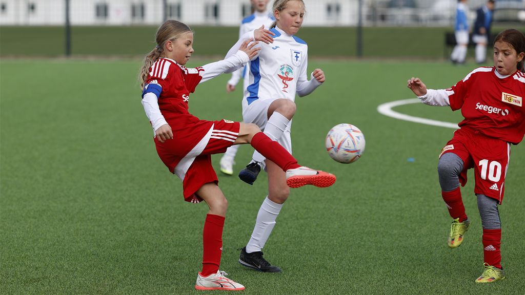 Drei Mädchen spielen Fußball auf einem grünen Rasenplatz, eine Spielerin hebt den Fuß zum Schuss.