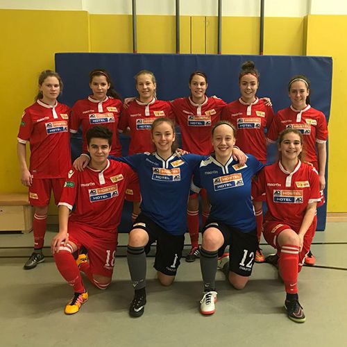 Eine Gruppe von zehn Frauen in rot-blauen Fußballtrikots steht und sitzt auf dem Boden in einer Sporthalle.