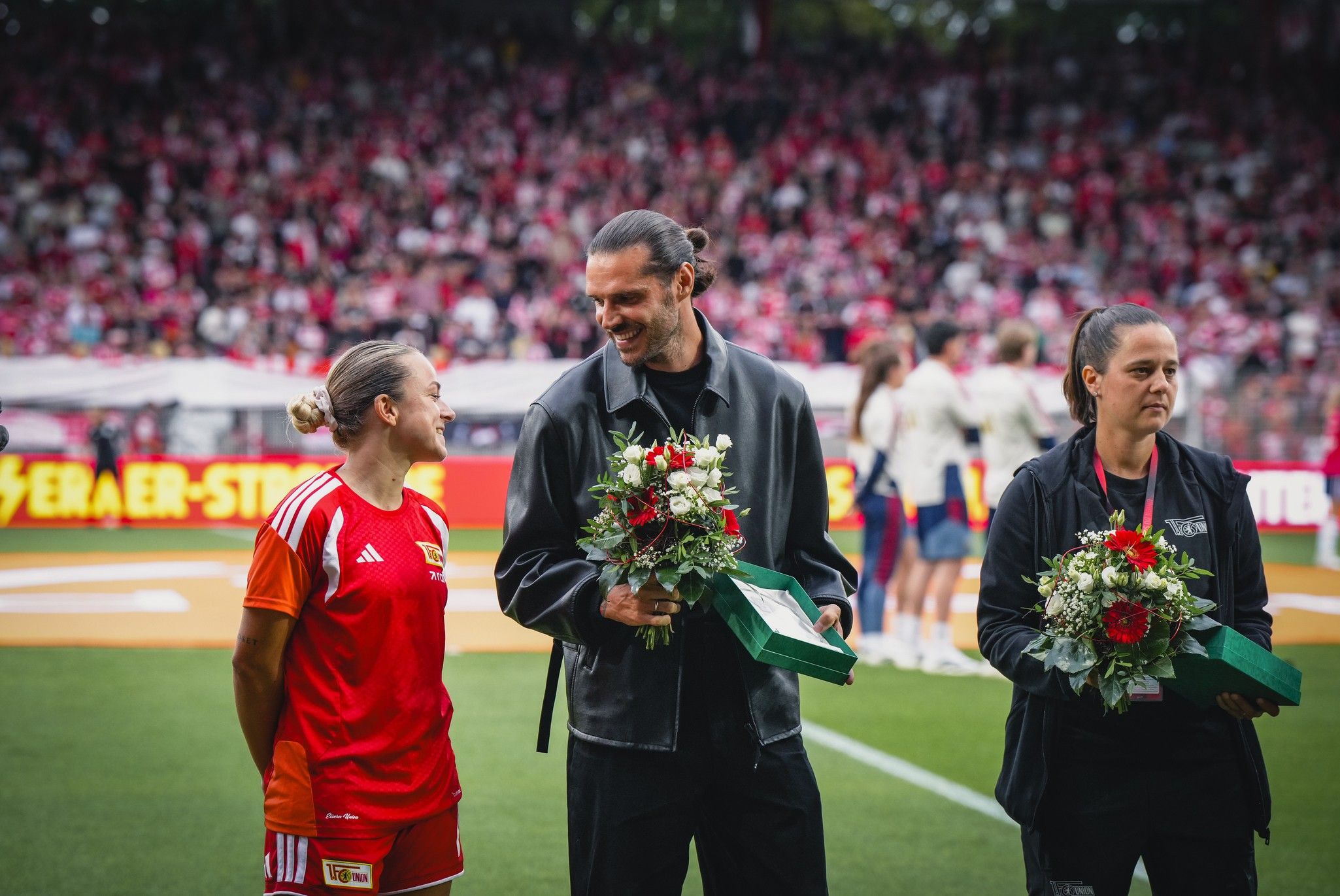 Eine Spielerin in Sportkleidung und ein Mann im schwarzen Outfit stehen lächelnd auf dem Platz, während eine Frau neben ihnen Blumen hält.