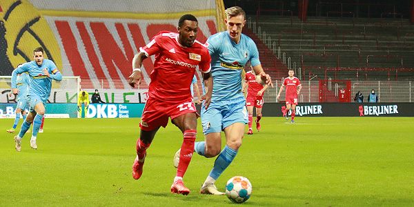 Zwei Fußballspieler in roten und blauen Trikots kämpfen um den Ball auf dem Spielfeld. Im Hintergrund sind leere Stadionplätze.