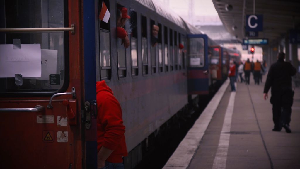 Zug am Bahnhof mit Passagieren, einige Köpfe sind zum Fenster herausgebeugt, unscharfer Hintergrund.