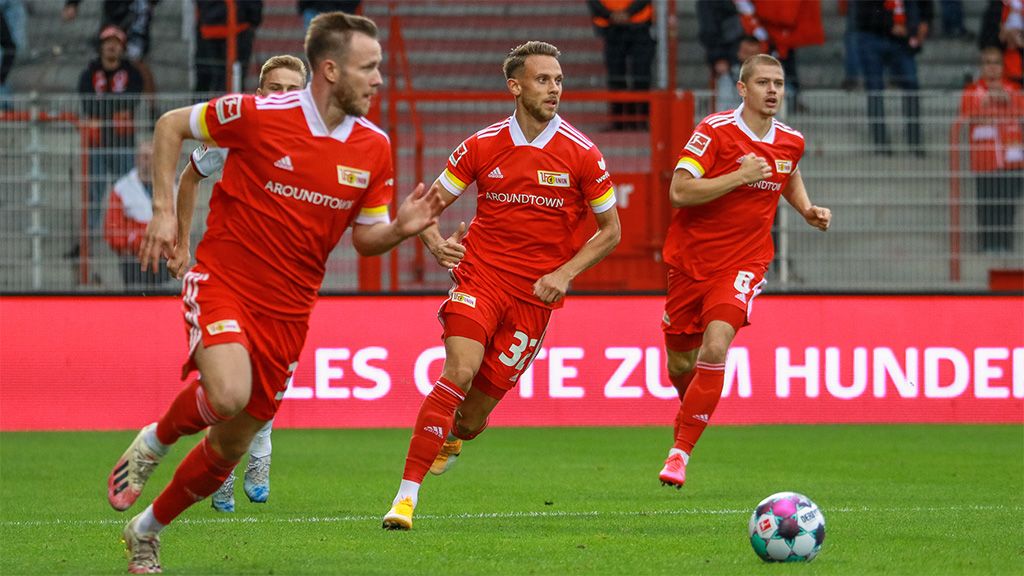 Drei Fußballspieler in roten Trikots laufen über das Spielfeld, während ein Ball im Vordergrund liegt.