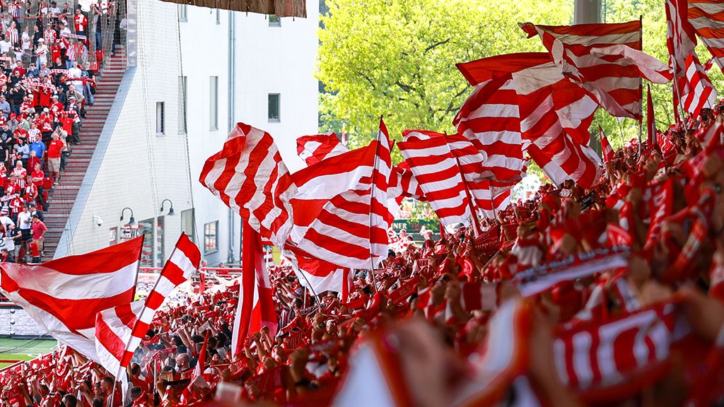 Eine jubelnde Menschenmenge in einem Stadion, die rote und weiße Fahnen schwenkt.