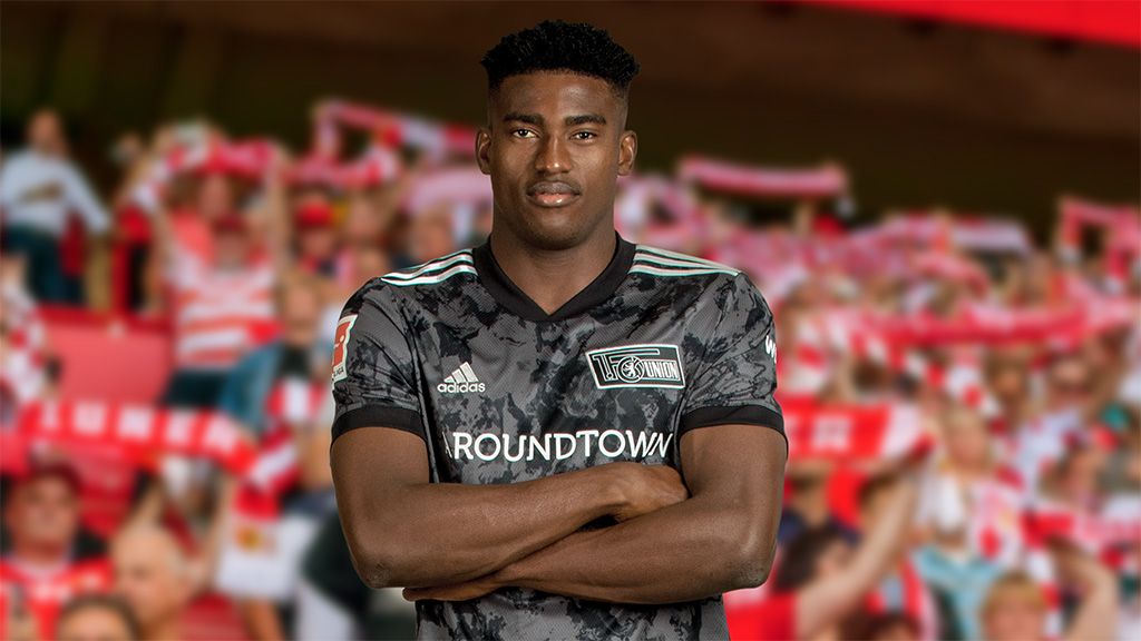 A young man dressed in sportswear stands with crossed arms in front of a cheering crowd in a stadium.