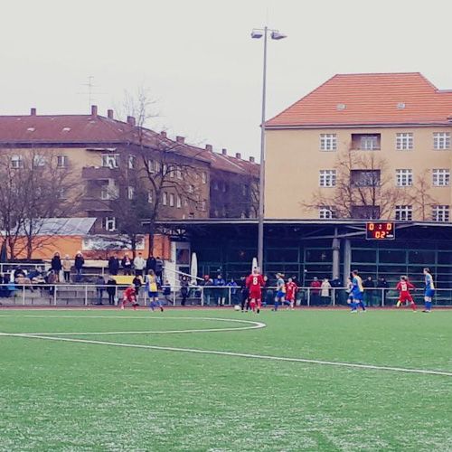 Ein Fußballspiel auf Kunstrasen. Die Spieler tragen rote und blaue Trikots. Im Hintergrund sind Zuschauer.