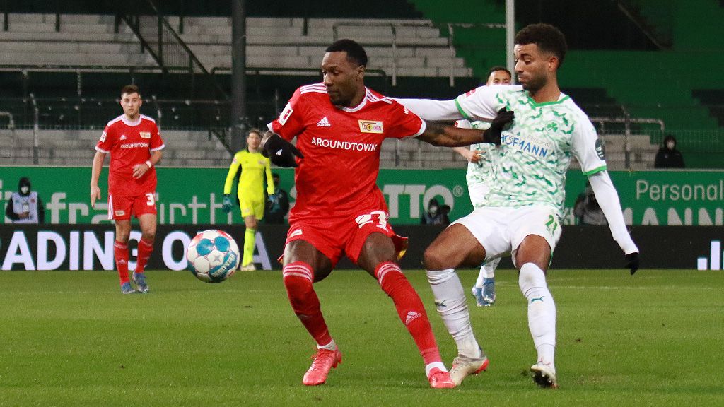 Two soccer players in red and green jerseys are battling for the ball on a field. In the background, there are more players and spectators.
