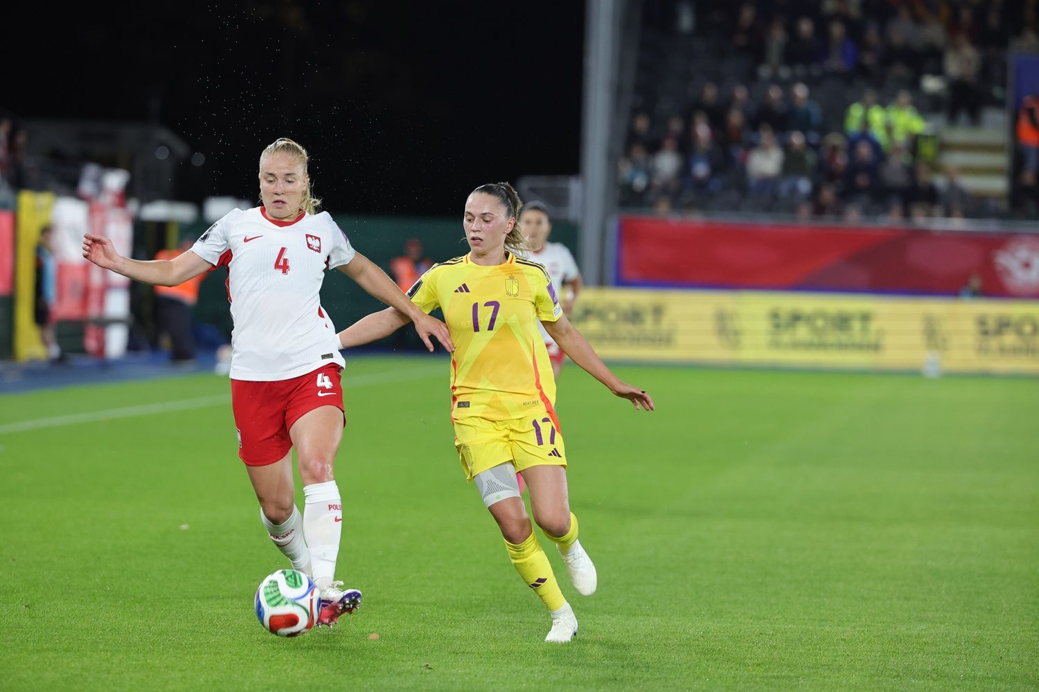 Zwei Frauenfußballspielerinnen im Wettkampf um den Ball auf einem Sportplatz, Zuschauer im Hintergrund.