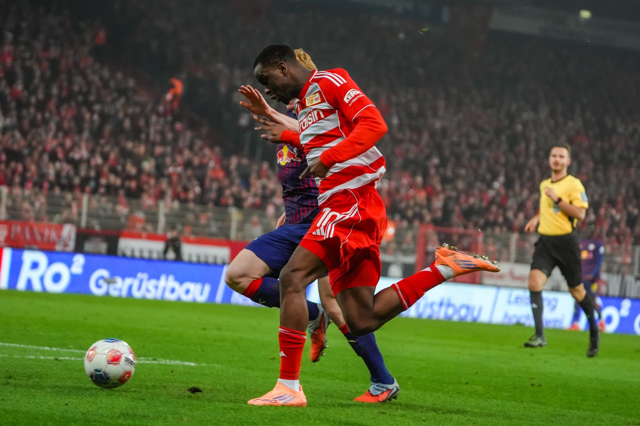 Fußballspiel mit einem Spieler in roter Kleidung, der den Ball beim Sprinten zu einem anderen Spieler in blauer Kleidung spielt.