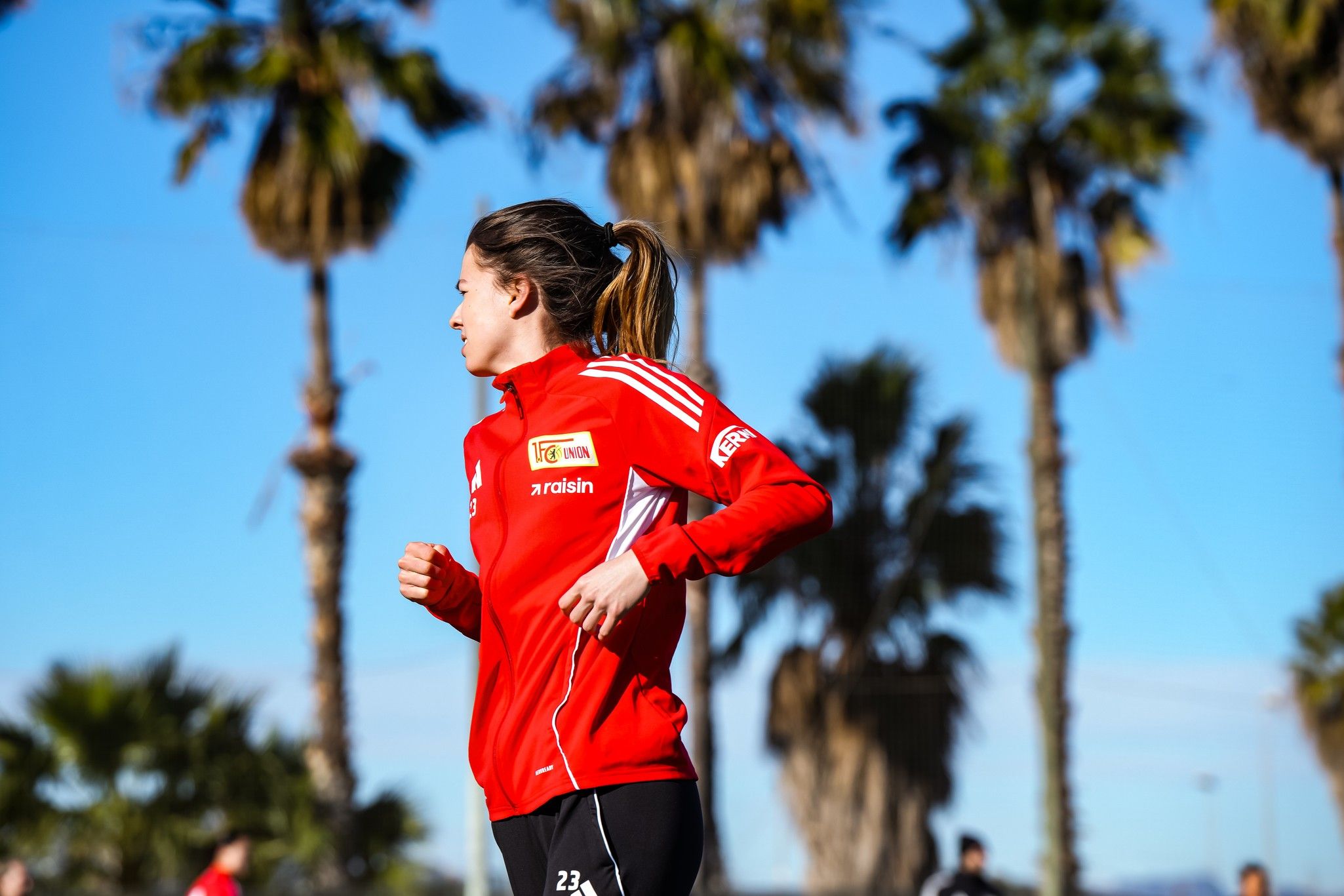 Eine Person in roter Sportbekleidung joggt vor Palmen und blauem Himmel.