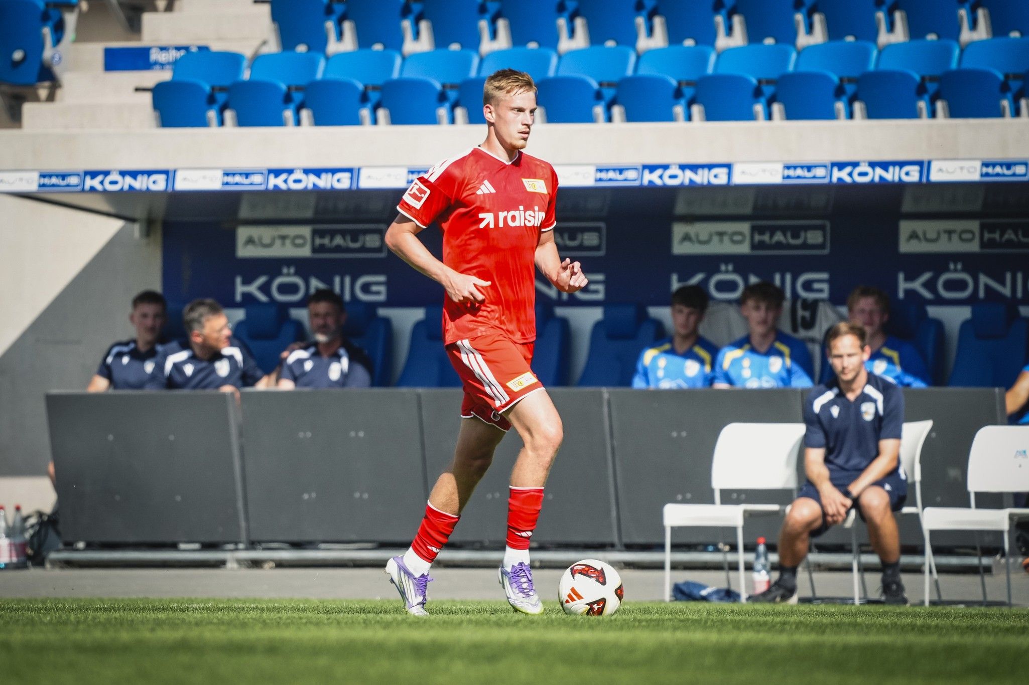 Ein Fußballspieler in roter Uniform läuft auf dem Spielfeld, während Zuschauer auf blauen Rängen sitzen.