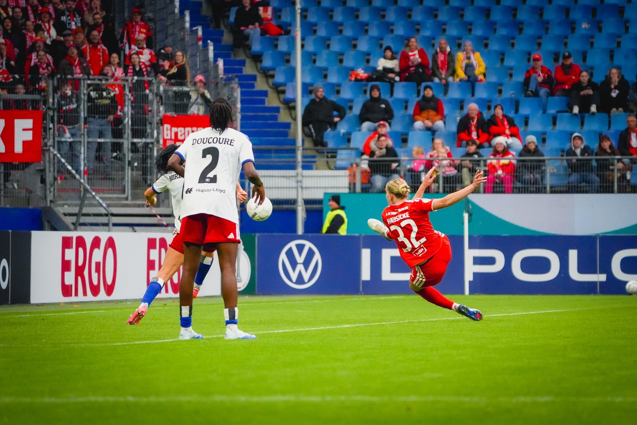 Eine Fußballspielszene mit einem Spieler in rotem Trikot, der einen Schuss aufs Tor abgibt, während ein Verteidiger versucht zu blocken.