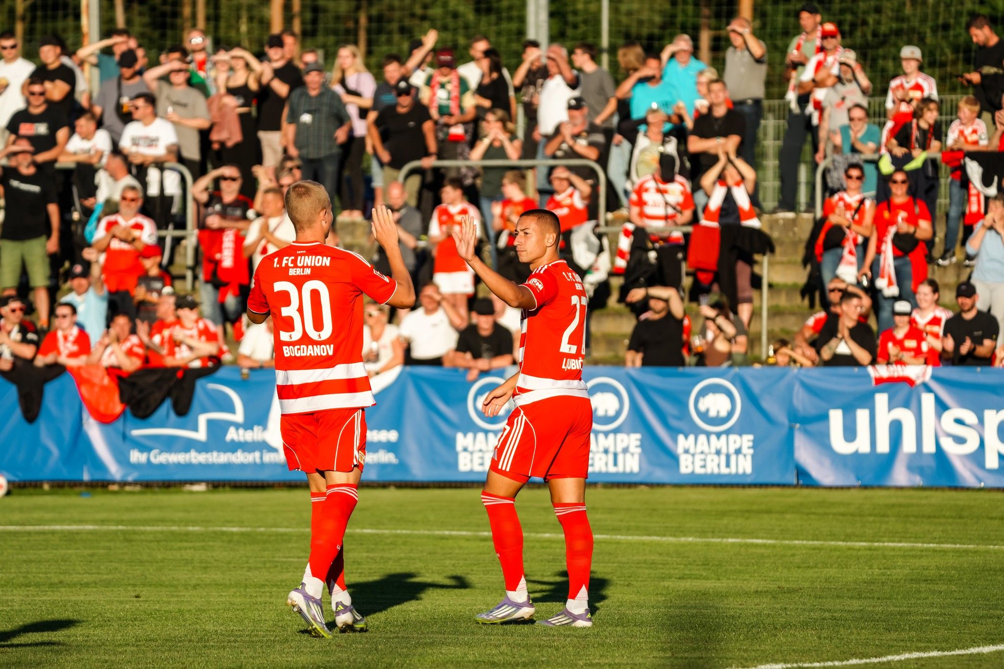 Zwei Fußballspieler in roten Trikots feiern vor jubelndem Publikum auf einem Sportplatz.