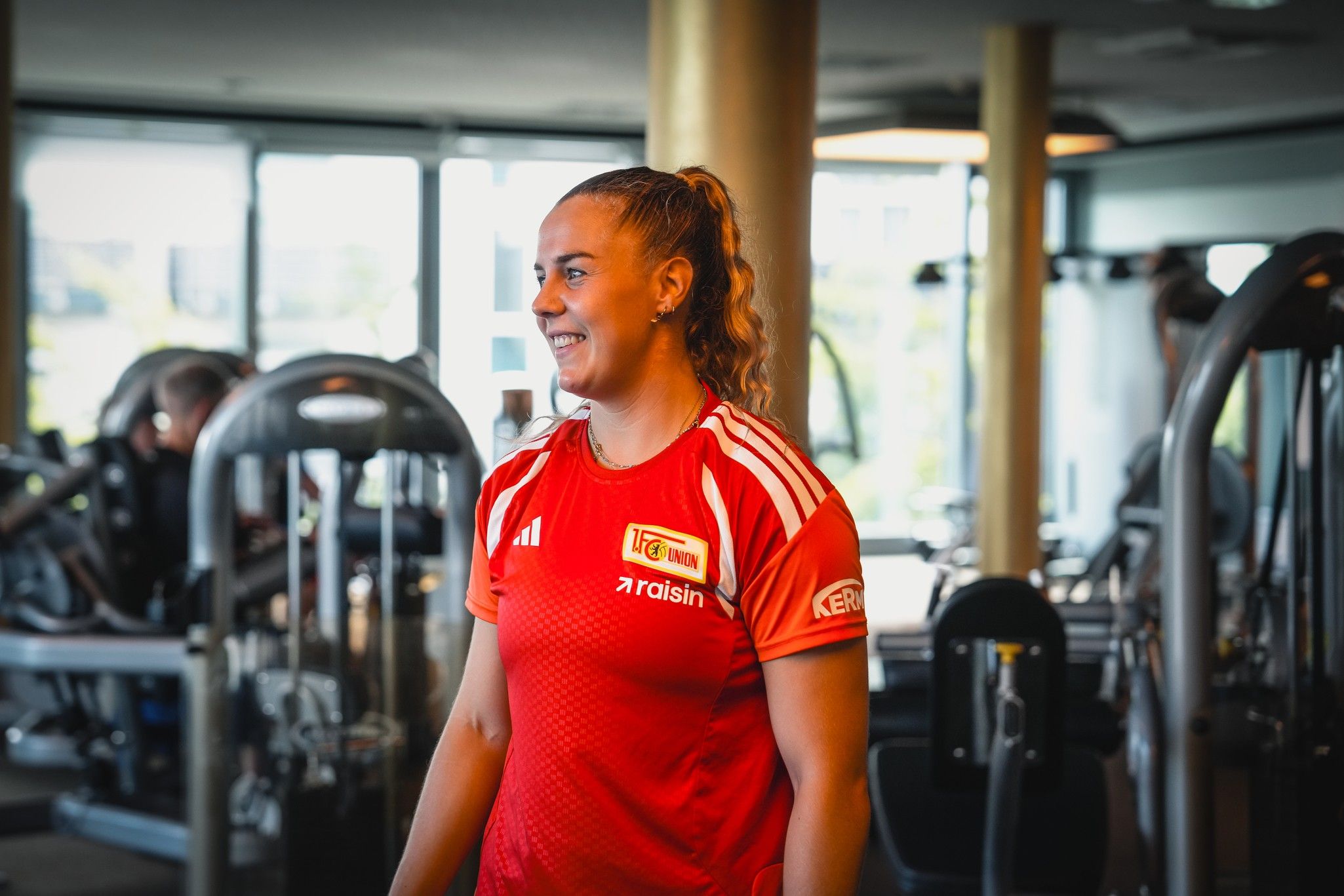 Eine sportliche Frau in einem roten Trikot lächelt in einem Fitnessstudio, umgeben von Trainingsgeräten.