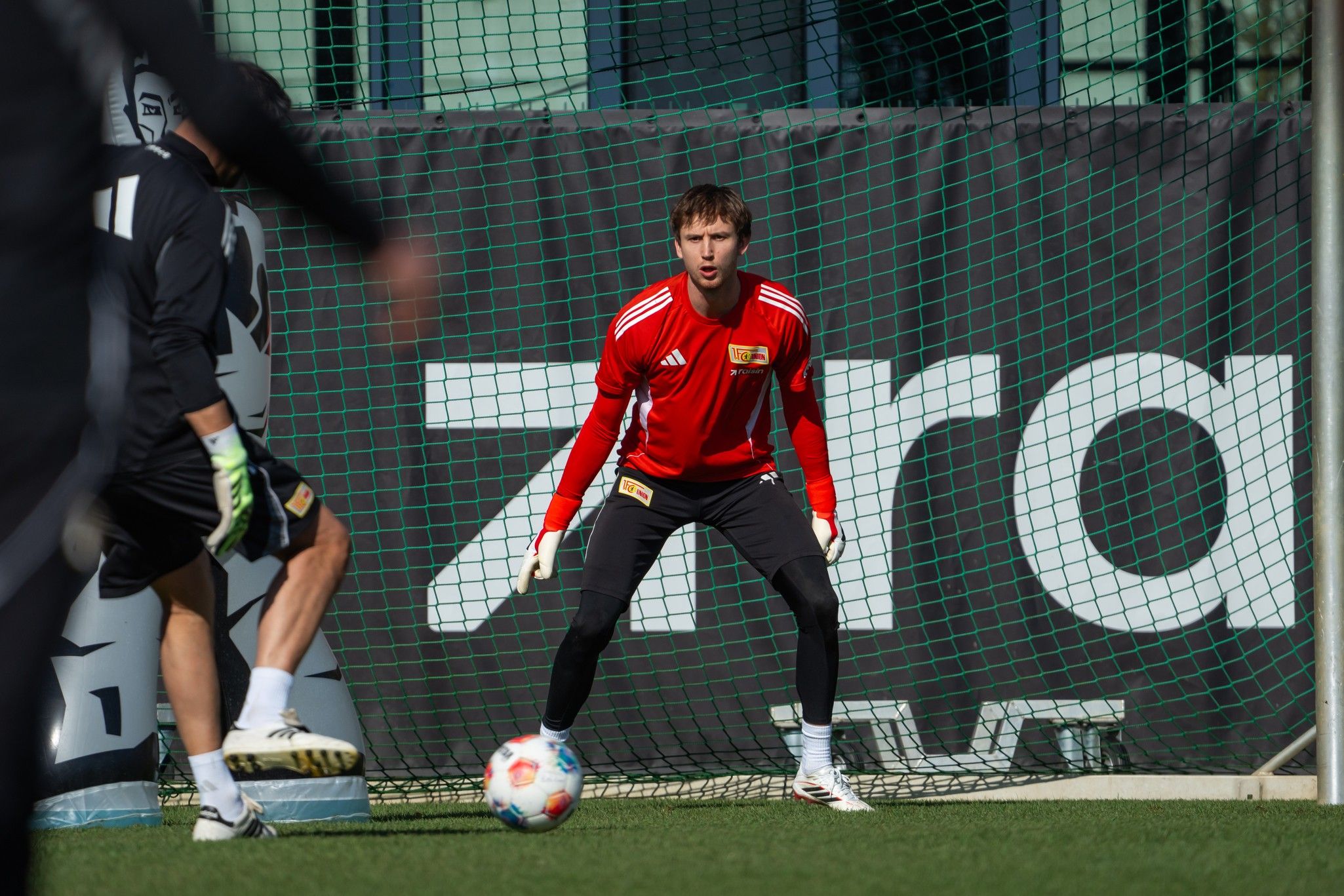 Ein Fußballtorwart in rotem Trikot steht im Tor, während ein Mitspieler einen Ball schießt. Hintergrund mit Netz und Werbebanner.