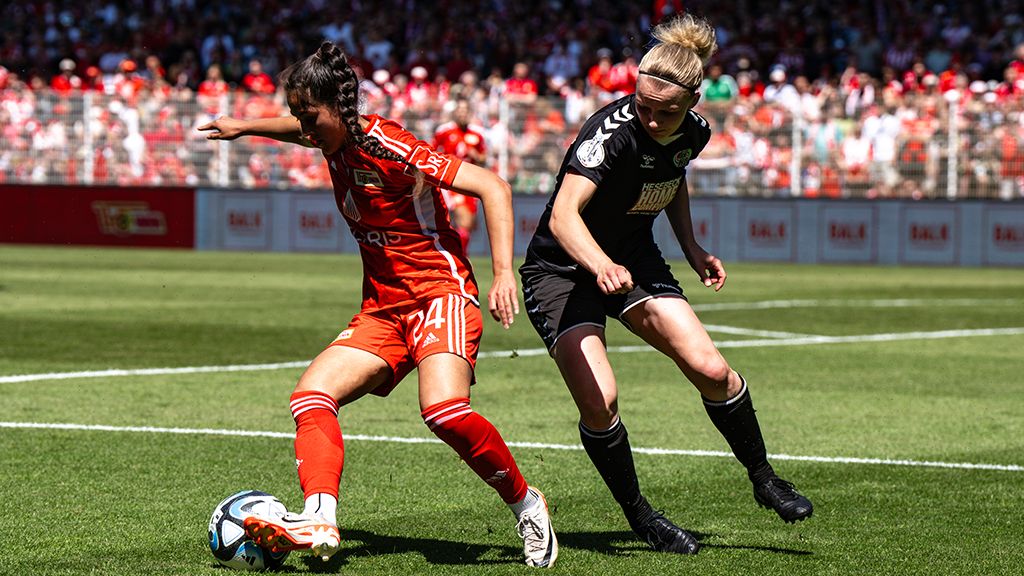Zwei Fußballspielerinnen, eine in roten und eine in schwarzen Trikots, kämpfen um den Ball auf einem Spielfeld.