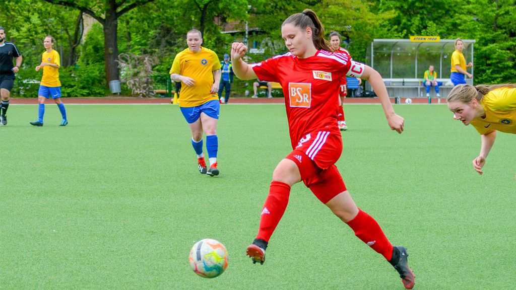 Eine Fußballspielerin in roter Uniform schießt den Ball, während andere Spielerinnen in gelben Trikots im Hintergrund stehen.