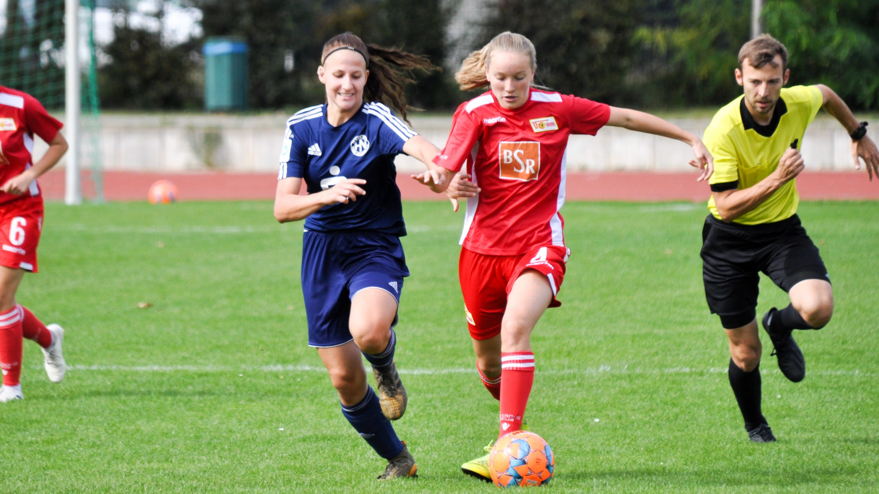 Zwei Fußballspielerinnen im Wettlauf um den Ball, während ein Schiedsrichter im Hintergrund das Spiel beobachtet.