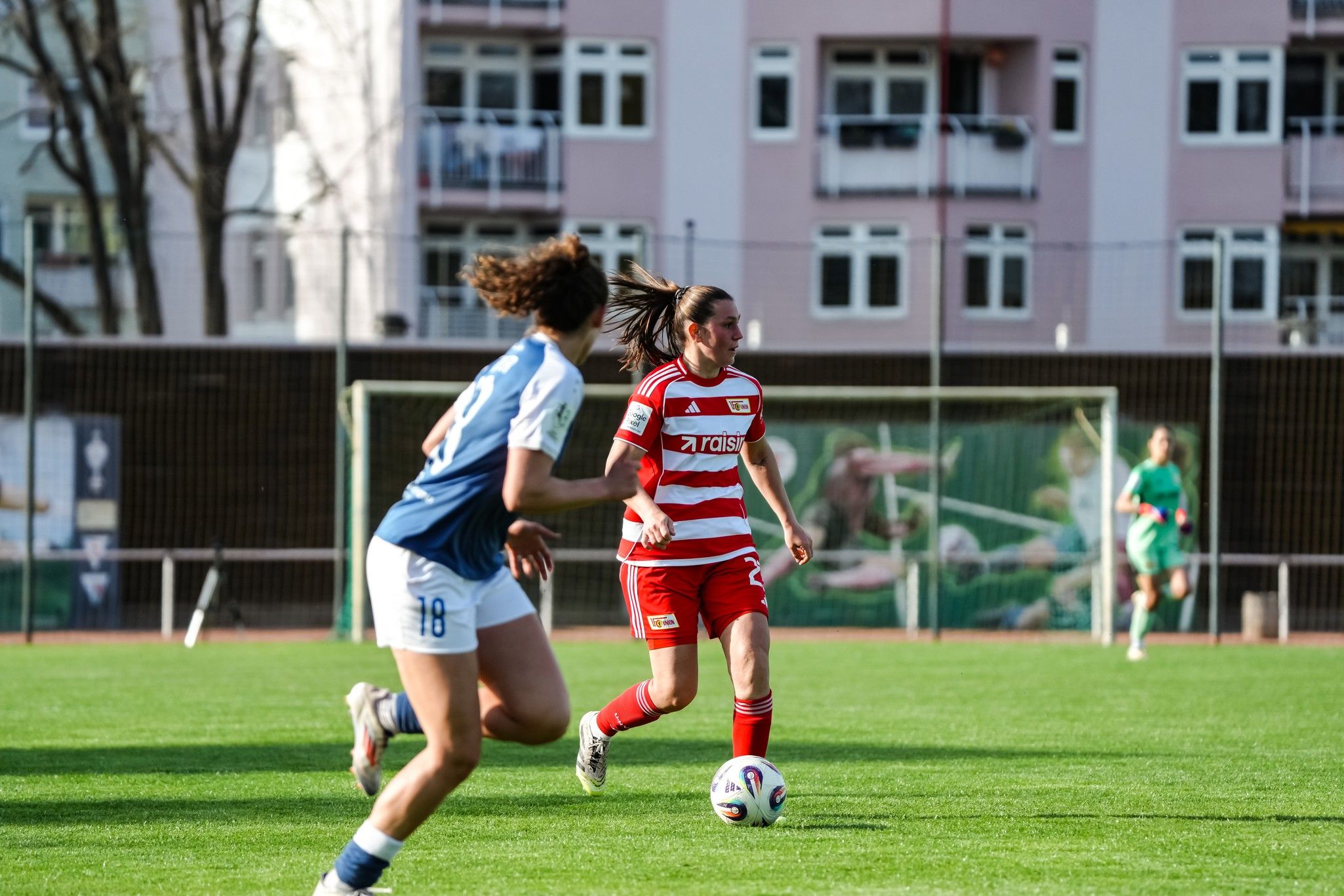 Eine Fußballspielerin in roter und weißer Uniform dribbelt den Ball auf dem Feld, während eine Gegnerin in blauer Uniform zusieht.