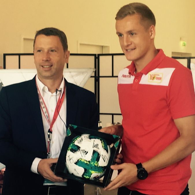 Zwei Männer stehen nebeneinander und halten einen signierten Fußball in einer Präsentationsumgebung.