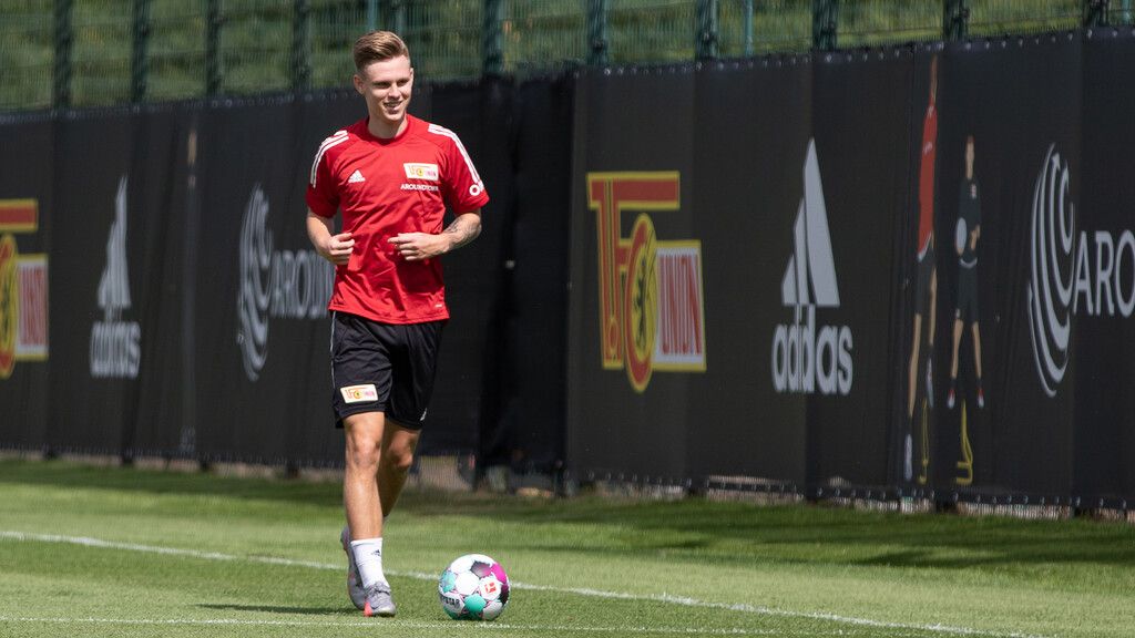 Ein Fußballspieler in rotem Trikot joggt auf einem Trainingsplatz, umgeben von Werbebannern und grüner Rasenfläche.