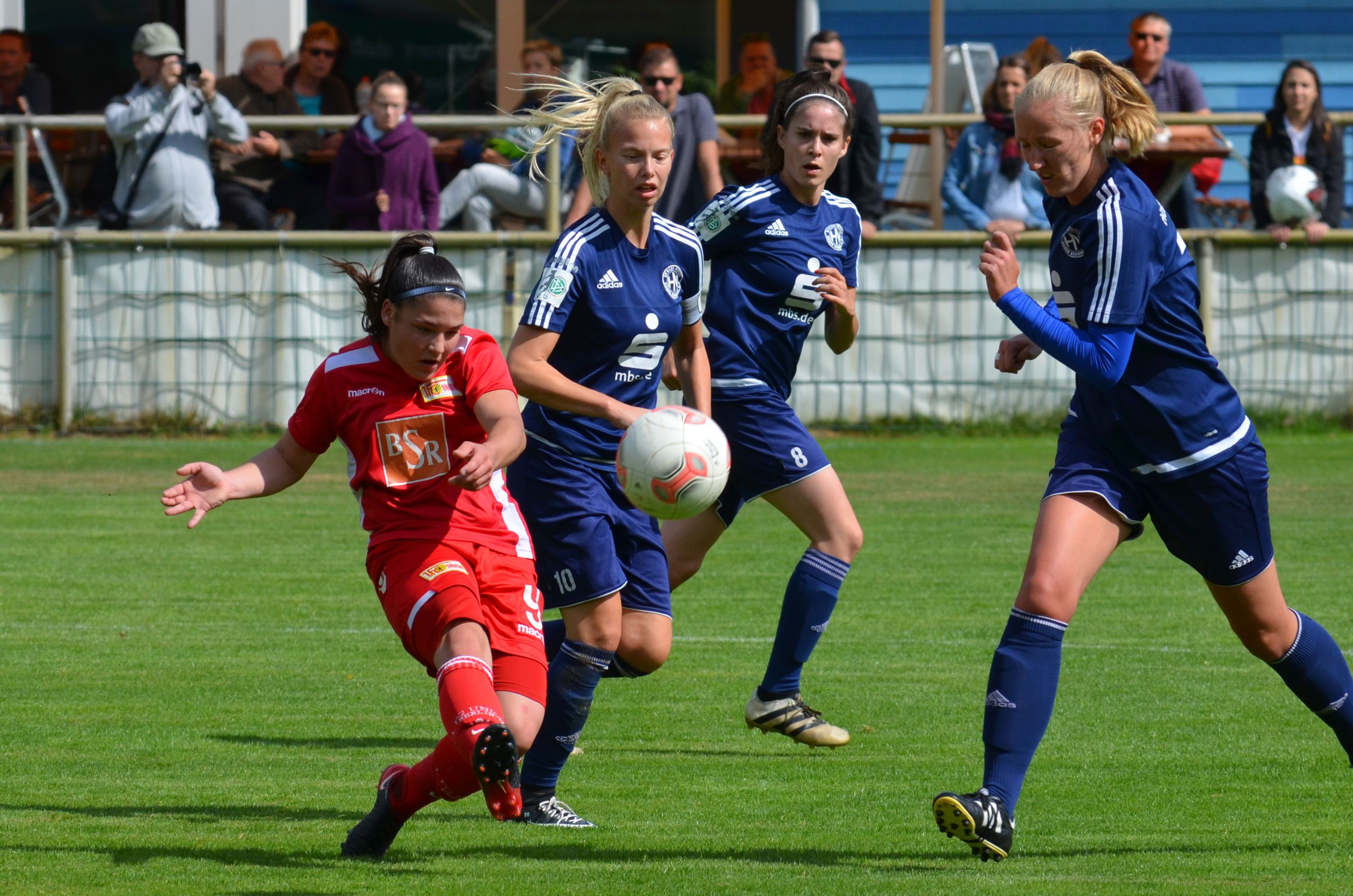 Eine Spielerin hat ein rotes Trikot an. Sie tritt den Ball. Drei Gegnerinnen in blauen Trikots versuchen, sie zu stoppen. Im Hintergrund sind Zuschauer.