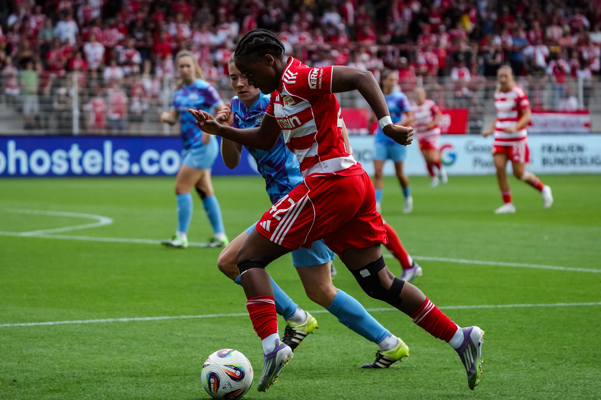 Spielerin in rote und weiße Kleidung dribbelt einen Fußball, während eine verteidigende Spielerin in blau versucht, sie zu verfolgen.