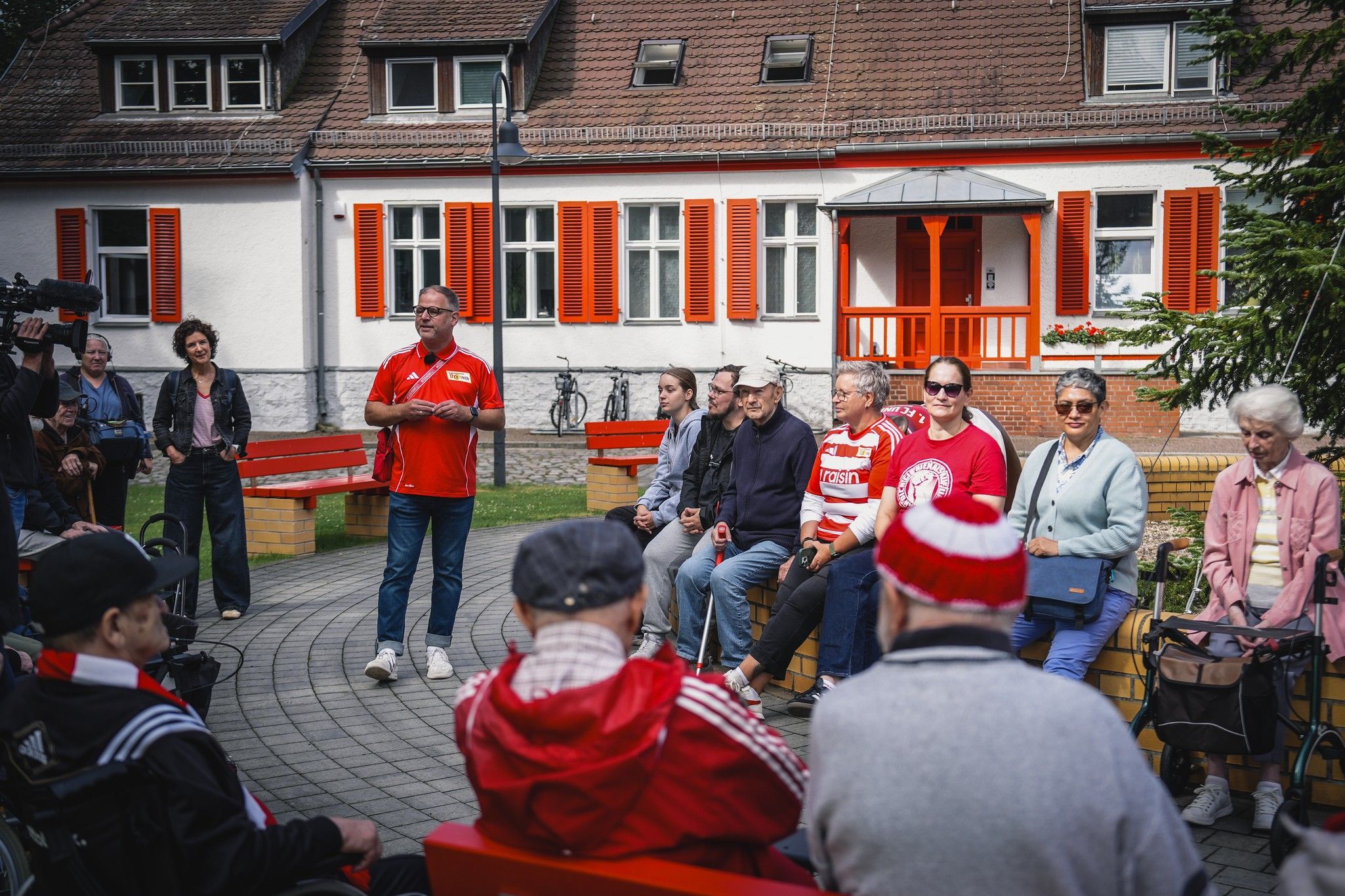Eine Gruppe von Menschen sitzt auf Bänken in einem Innenhof, während ein Mann in rotem Shirt spricht. Hintergründe mit Fenstern und Bäumen.