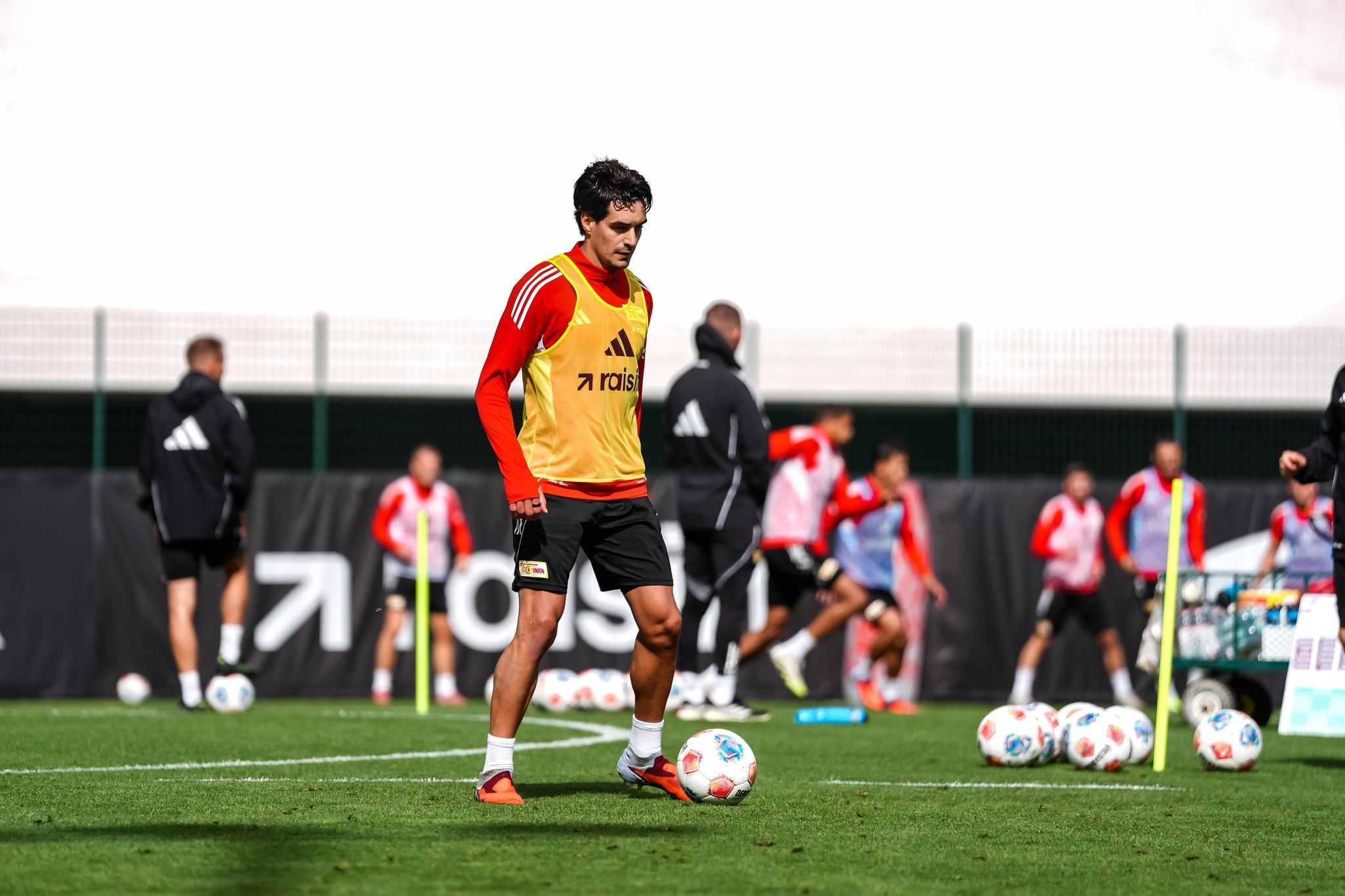 Fußballspieler in Trainingskleidung übt auf dem Platz, während andere Spieler im Hintergrund trainieren.