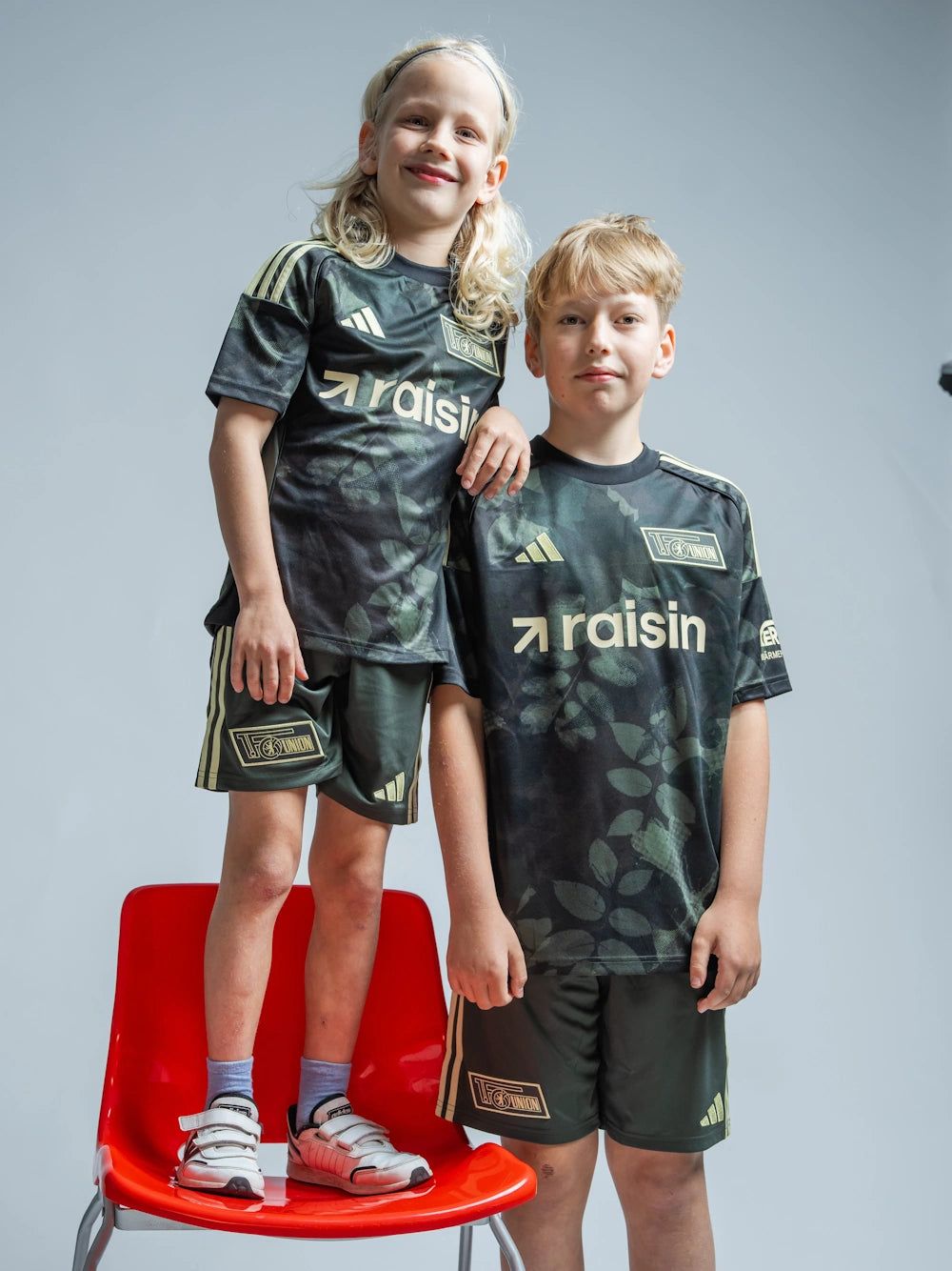 Ein Junge und ein Mädchen tragen sportliche Trikots mit dem Schriftzug "raisin" auf einem roten Stuhl vor einem grauen Hintergrund.