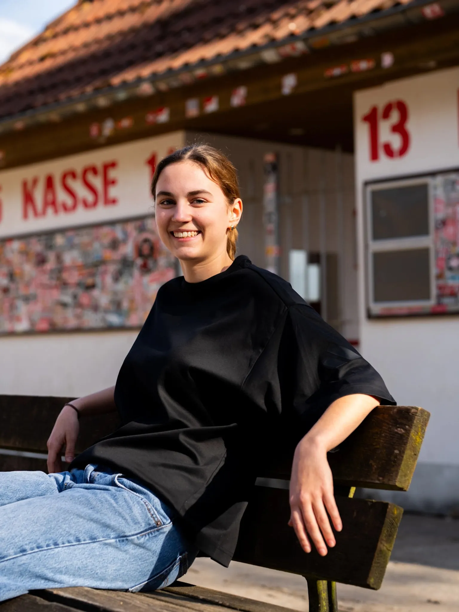 Junge Frau mit kurzer brauner Haarfrisur sitzt auf einer Bank und lächelt, im Hintergrund ein Gebäude mit der Aufschrift „KASSE“.
