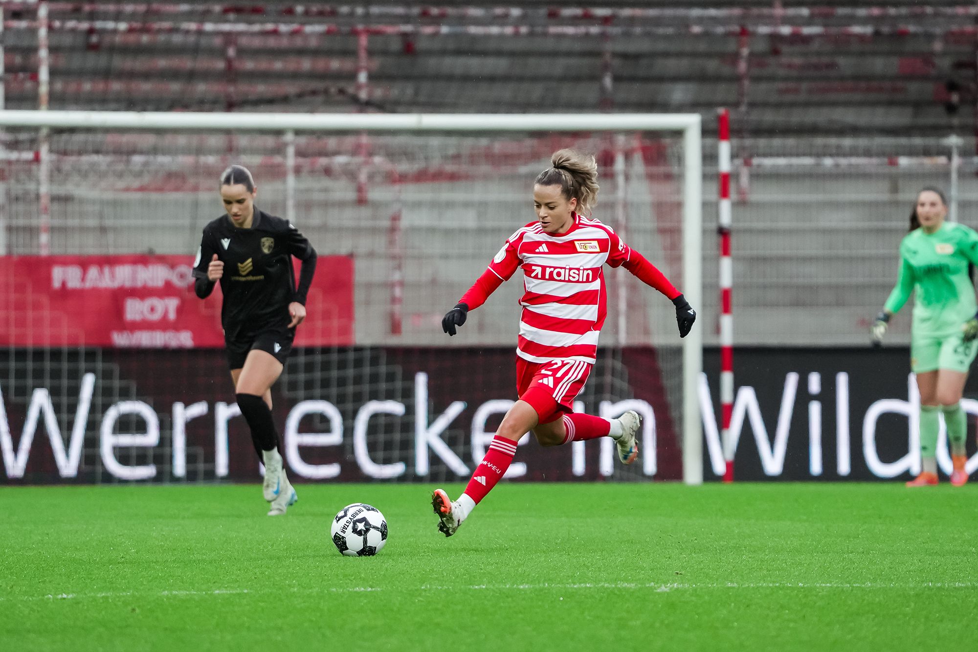 Eine Fußballspielerin in rot-weißer Uniform tritt den Ball, während zwei Gegenspielerinnen im Hintergrund stehen.