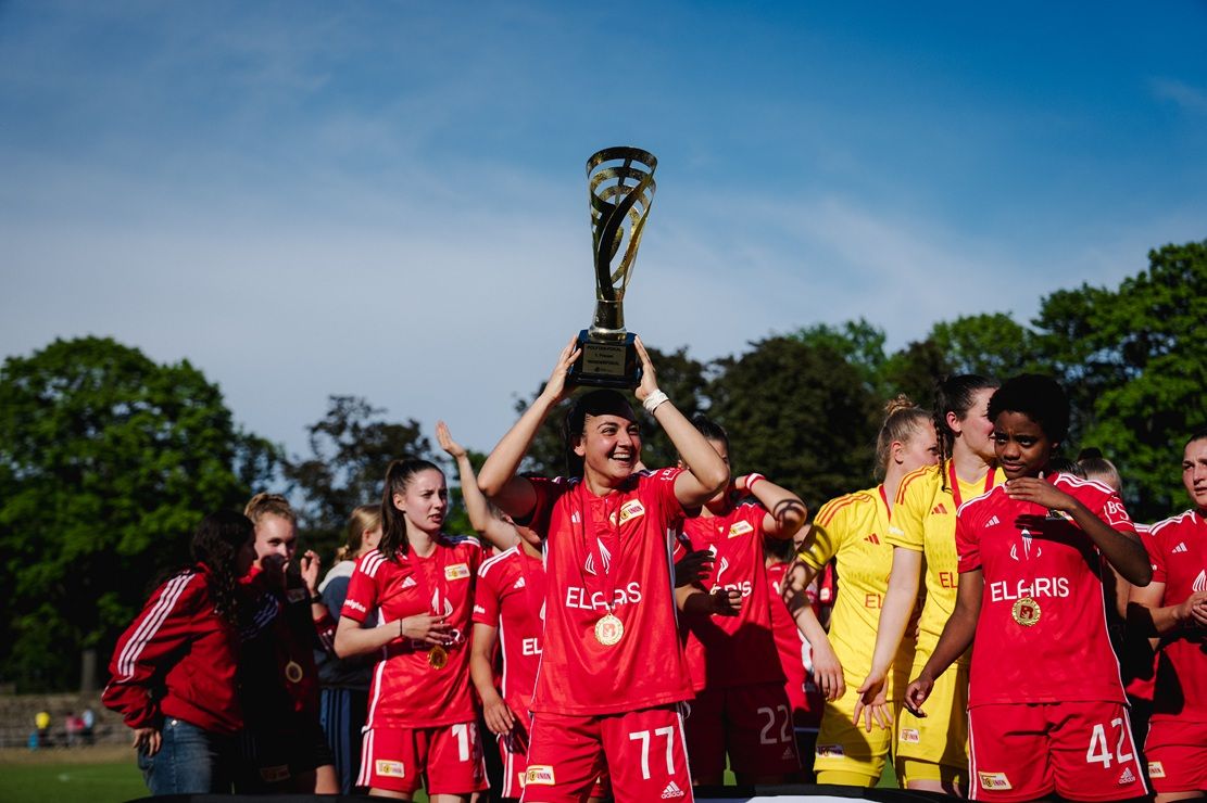 Eine Fußballmannschaft in roten Trikots feiert den Sieg mit einem Pokal unter blauem Himmel.