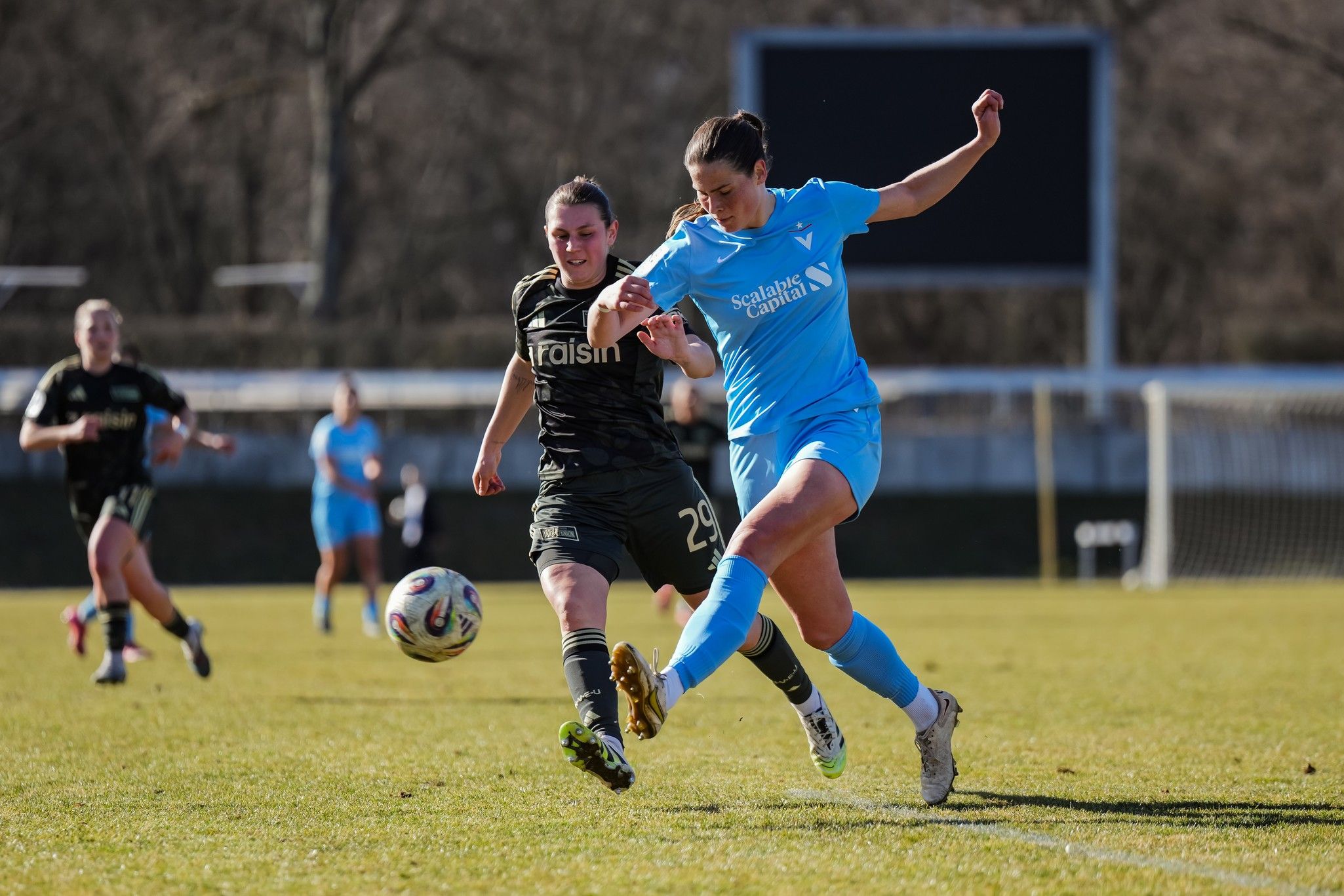 Zwei Fußballspielerinnen im Wettkampf um den Ball auf einem Rasenplatz, im Hintergrund weitere Spielerinnen.
